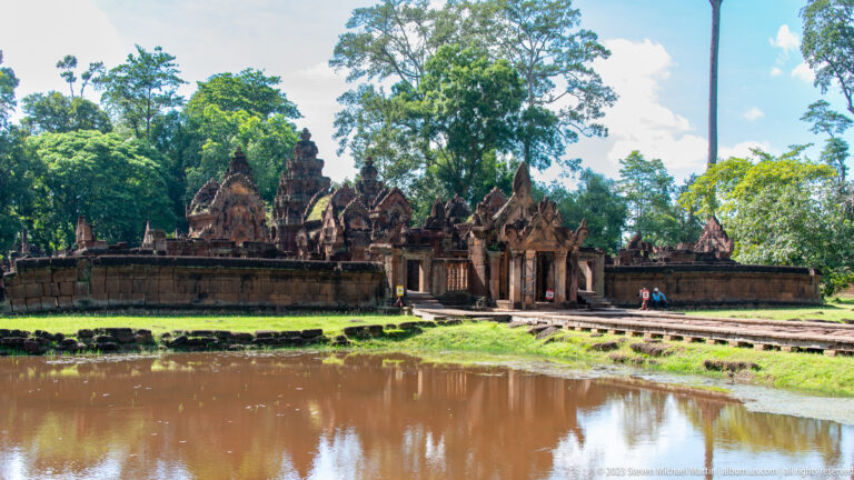 Wide view across a moat toward Banteay Srei's enclosure wall, entrance gate, and temple roofs.