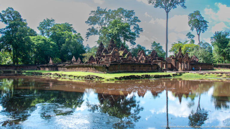 Temple enclosure and towers reflected in the broad moat beneath tall trees and blue sky.
