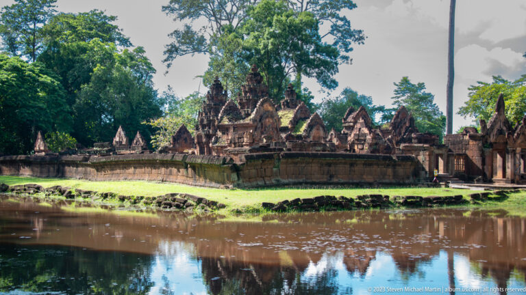 Long sandstone enclosure wall and temple buildings viewed across the moat with reflections in the water.