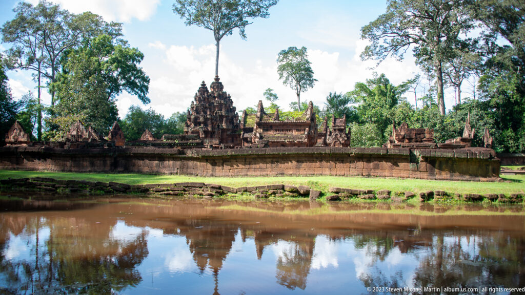 Broad view of Banteay Srei across the moat, with the central tower and trees reflected in the water.