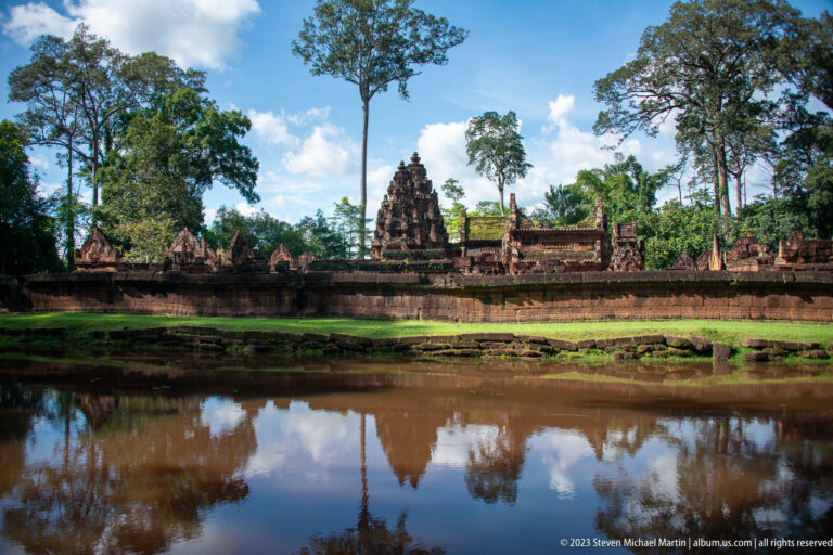 Wide view of Banteay Srei's central sanctuary tower and enclosure reflected in still water beneath tall trees.