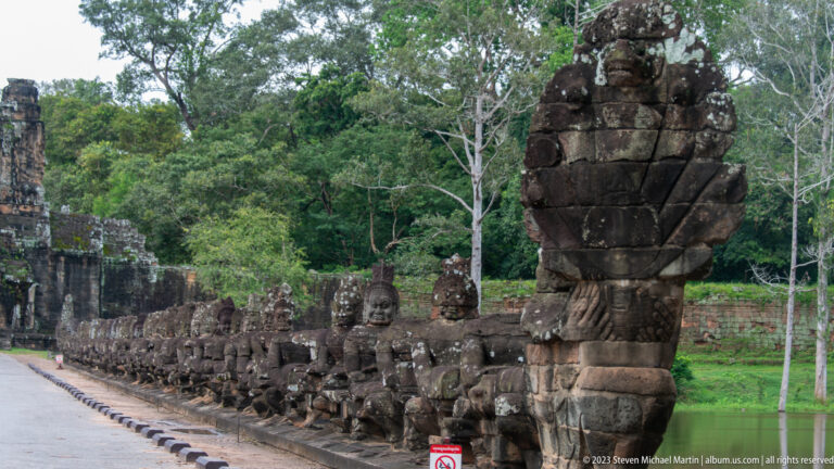 Stone guardian figures holding a naga balustrade beside a moat, with a large serpent head in the foreground.