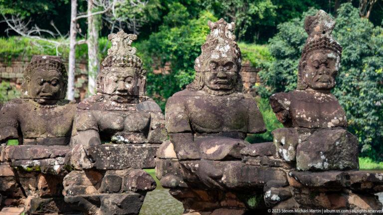 Four weathered seated stone guardians holding a naga balustrade in bright sunlight.