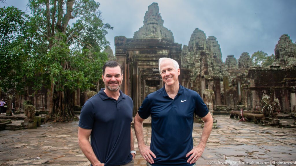 Two male visitors posing together in a Bayon courtyard with face towers behind them.