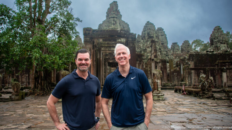 Closer portrait of two male visitors smiling in front of Bayon Temple's central towers.