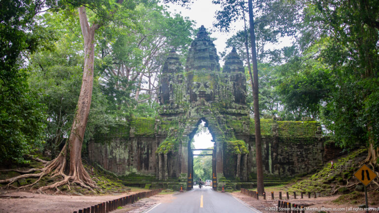 Road leading through a moss-covered face-tower gate surrounded by large trees at Angkor Thom.