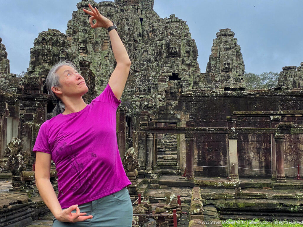 Female visitor in a magenta shirt posing with one arm raised in front of Bayon Temple.