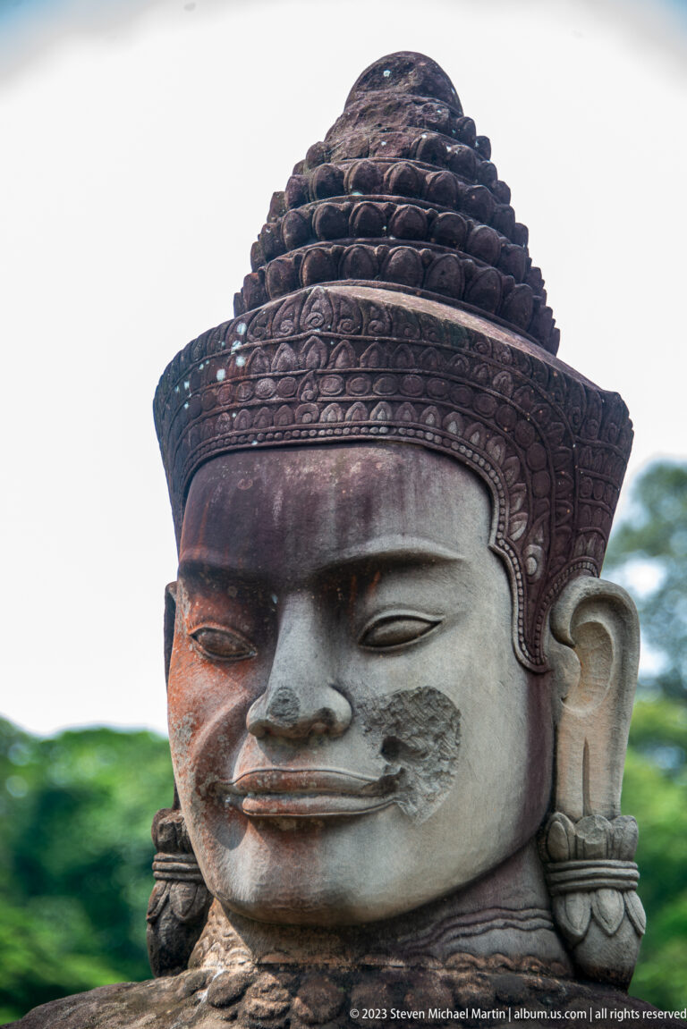 Close frontal portrait of a dark sandstone guardian face with damaged nose and pale cheek patch.