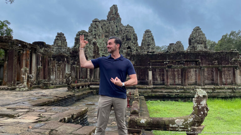 Male visitor raising one hand beside a weathered stone balustrade at Bayon Temple.