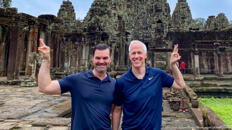 Two male visitors posing together with raised hands in front of Bayon Temple.
