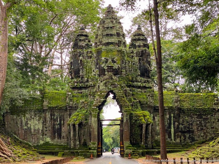 Frontal view of a moss-covered Angkor Thom face-tower gate framed by large trees.