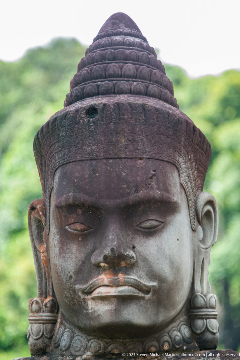 Centered close-up of a sandstone guardian face with broad patterned headdress and damaged nose.