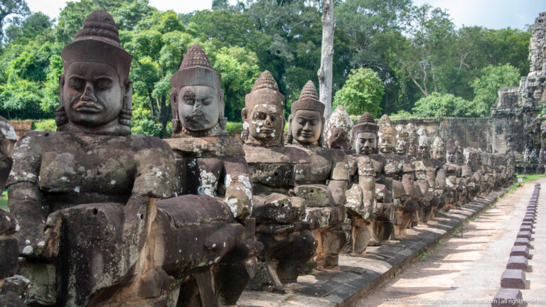 Diagonal row of seated stone guardians holding a naga beside the causeway road.