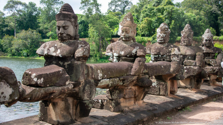 Side view of seated guardians gripping a stone naga balustrade beside water.