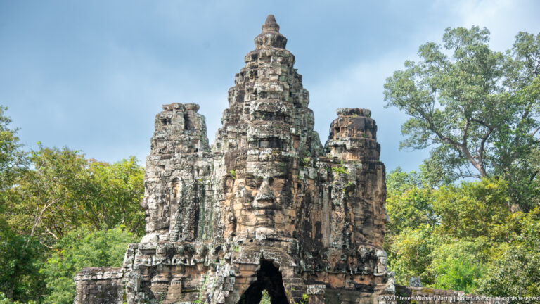 Weathered Angkor stone tower with a large carved face rising above trees.