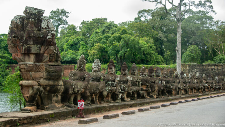 Long row of weathered stone guardian figures holding a naga beside the moat and road at Angkor Thom.