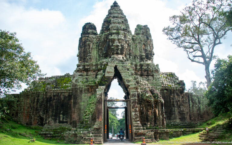 Wide frontal view of an Angkor Thom gate with people beneath the pointed arch.
