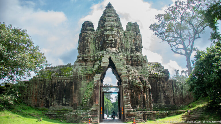 Angkor Thom gate framed by grass, tall trees, and open sky.