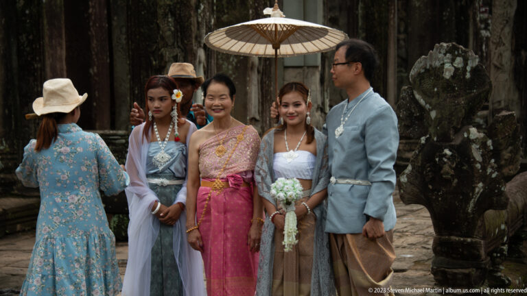 Five visitors in colorful formal clothing posing beneath a parasol at Bayon Temple.