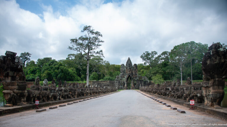 Straight road between two rows of stone guardian figures and naga balustrades leading toward an Angkor Thom gate.