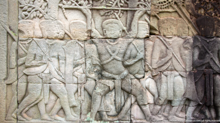 Khmer stone bas-relief of standing figures carrying long staffs or weapons.