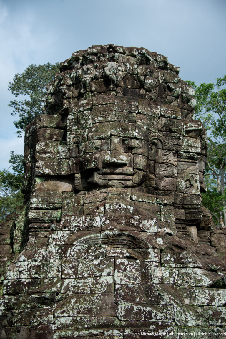 Weathered Bayon face tower with broken upper masonry against the sky.
