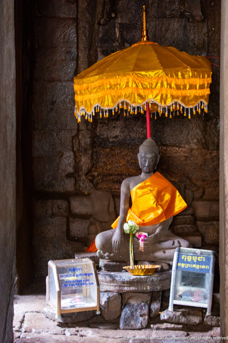 Seated Buddha in gold cloth beneath a golden parasol inside a dark stone chamber.