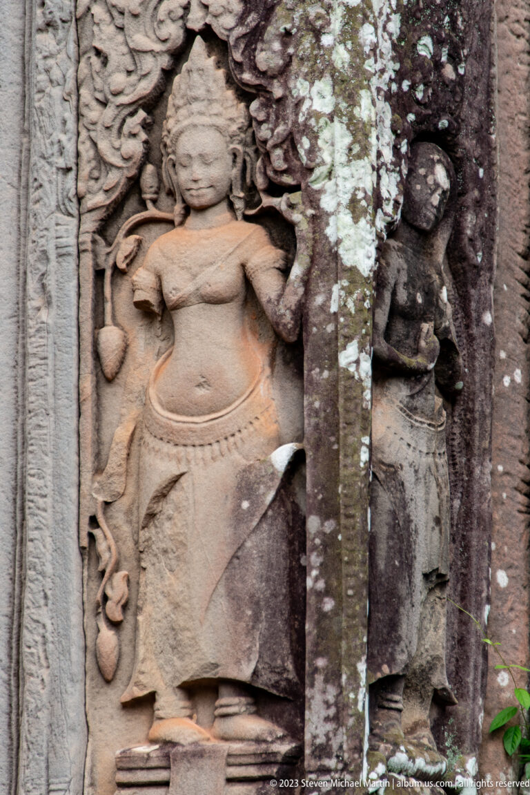 Standing female deity relief beside a darker damaged figure in a carved stone niche.