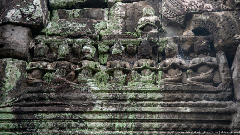 Weathered stone relief of small figures beneath ornament, with heavy green moss in the joints.