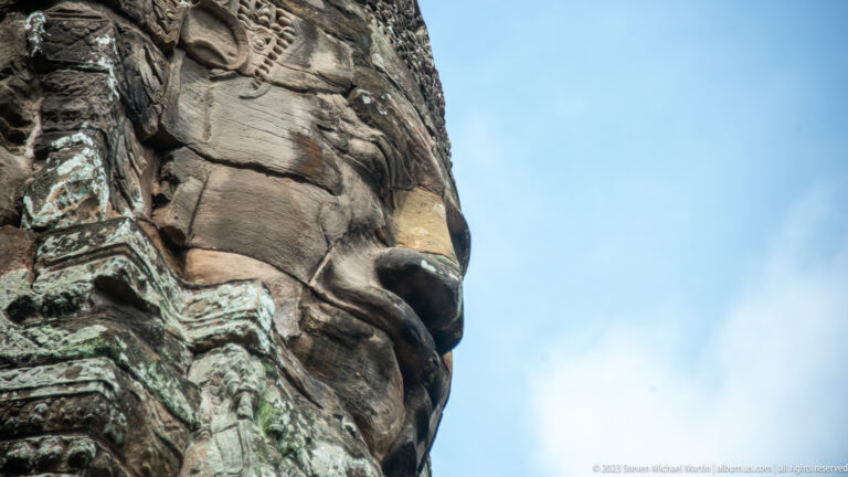 Side profile of a colossal Bayon face projecting against blue sky.
