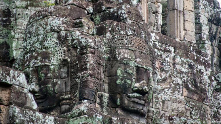Two carved Bayon faces on adjoining sides of a lichen-covered tower.