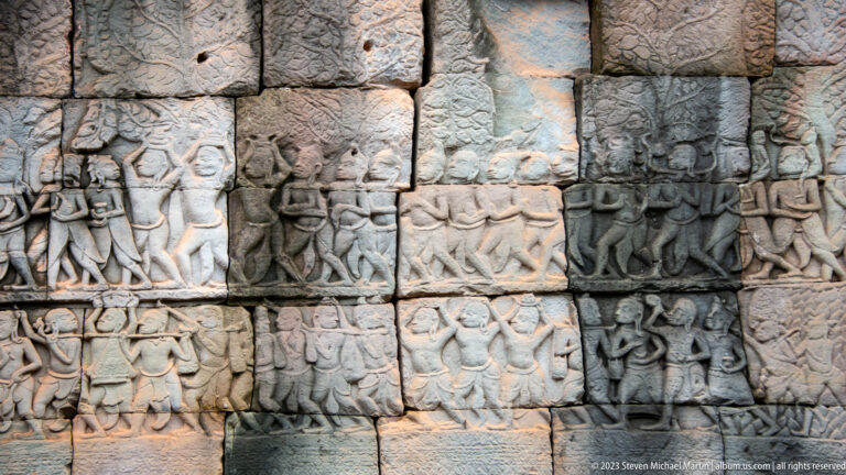 Multi-register Khmer relief of marching and crouching figures carrying long objects.