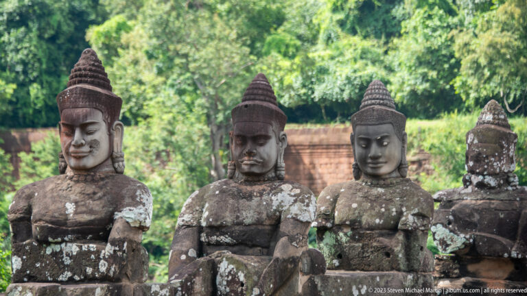 Close view of three weathered sandstone guardian figures with tall headdresses along an Angkor Thom causeway.