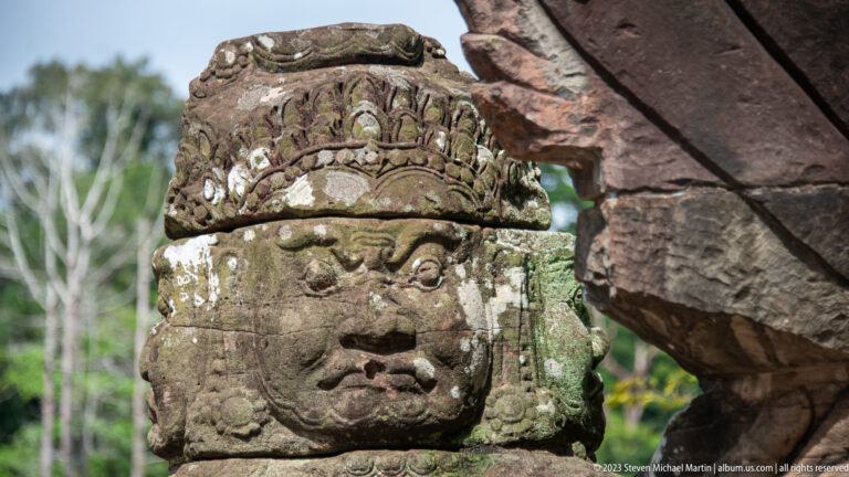 Close view of a weathered sandstone guardian head with ornate headdress and lichen-covered surface.