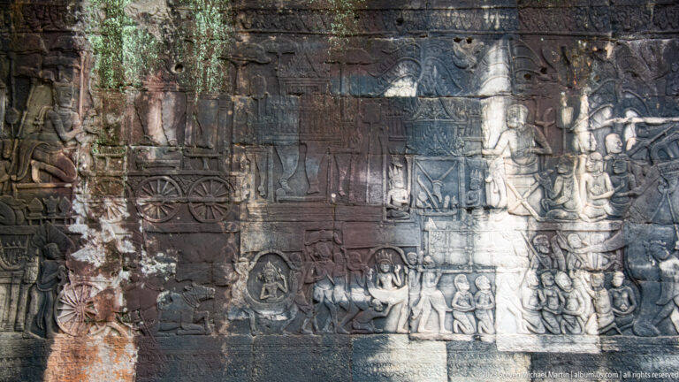 Wide Khmer bas-relief with chariots, processions, seated figures, and diagonal light.