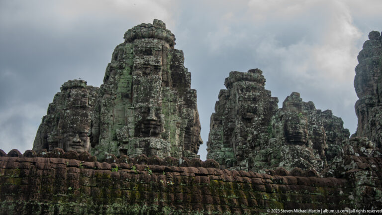 Low-angle view of Bayon Temple face towers above a moss-covered sandstone wall under dark clouds.