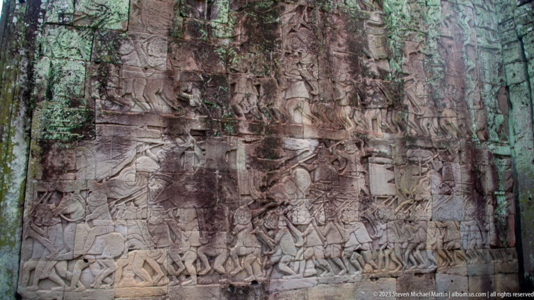 Weathered sandstone bas-relief with rows of standing, seated, and marching figures on a Bayon gallery wall.
