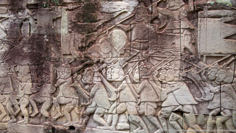 Sandstone bas-relief of a procession with a larger headdress-wearing figure, attendants, weapons, and standards.