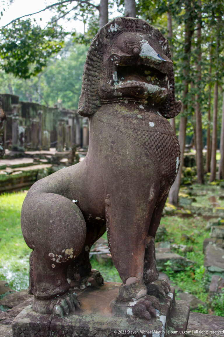 Weathered seated stone guardian lion with an open mouth and lichen-covered surface at Bayon Temple.
