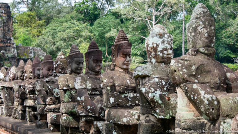 Long diagonal row of seated stone guardians holding a naga balustrade beside the Angkor Thom roadway.