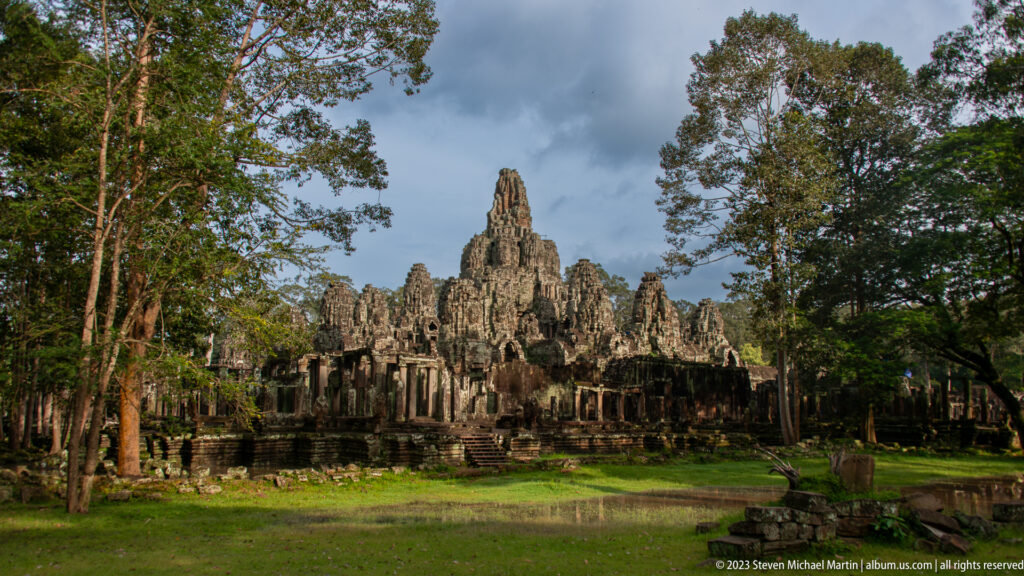 Wide view of Bayon Temple across grass with clustered towers and surrounding trees.