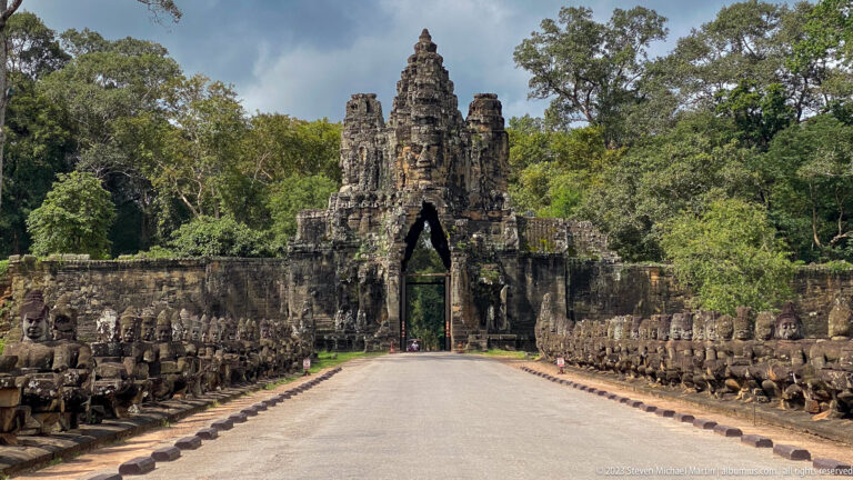 Broad centered view down the sculptural causeway toward an Angkor Thom face-tower gate.
