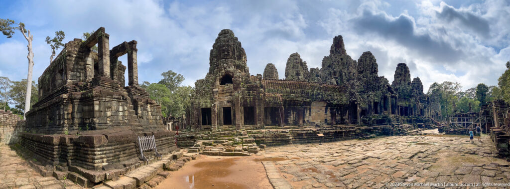 Oblique Bayon courtyard with broken paving, a ruined structure, galleries, and face towers.