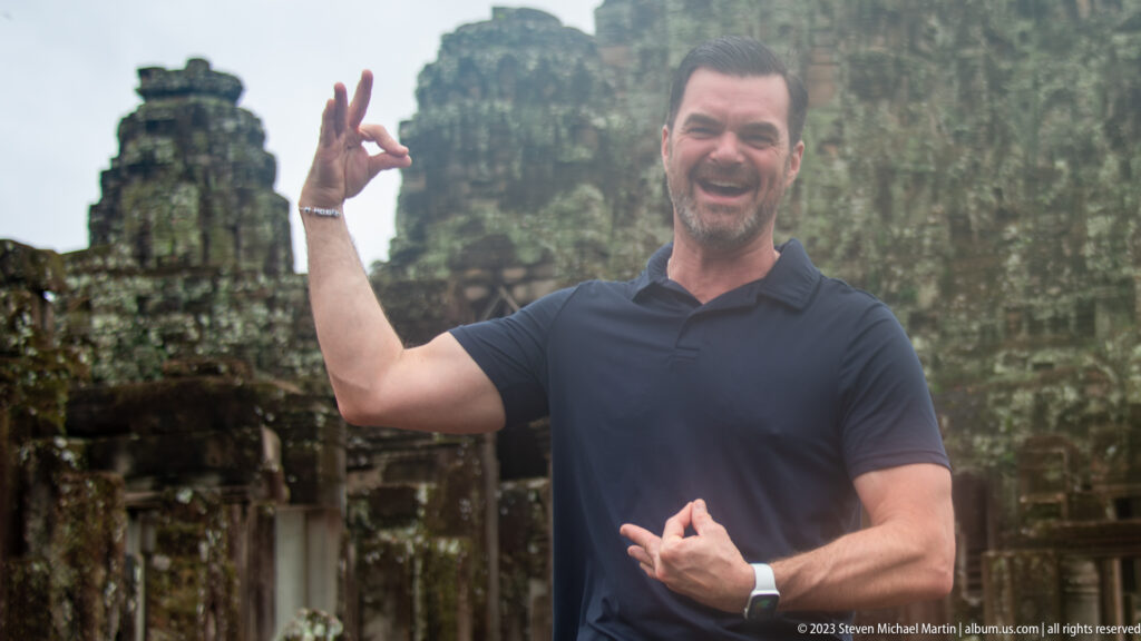 Smiling male visitor in a dark polo gesturing in front of Bayon Temple.