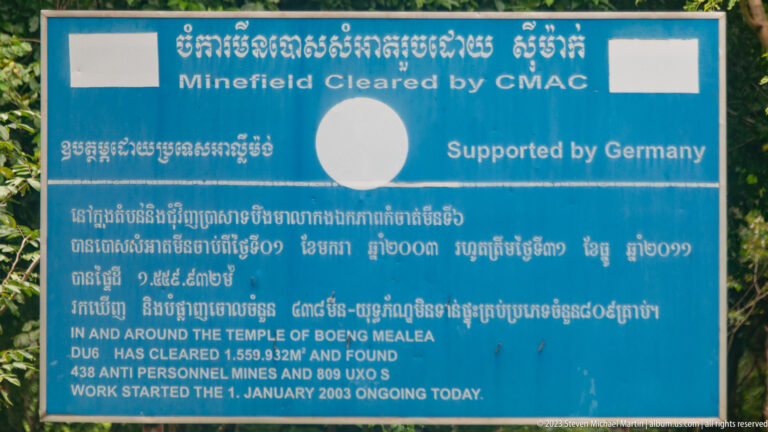 Blue CMAC sign with Khmer and English text about mine and unexploded-ordnance clearance at Beng Mealea.