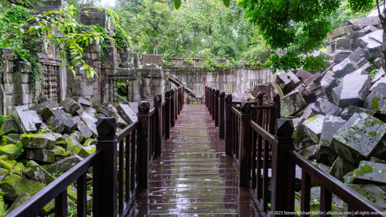 Wet wooden walkway runs between piles of collapsed sandstone blocks and forest vegetation at Beng Mealea.
