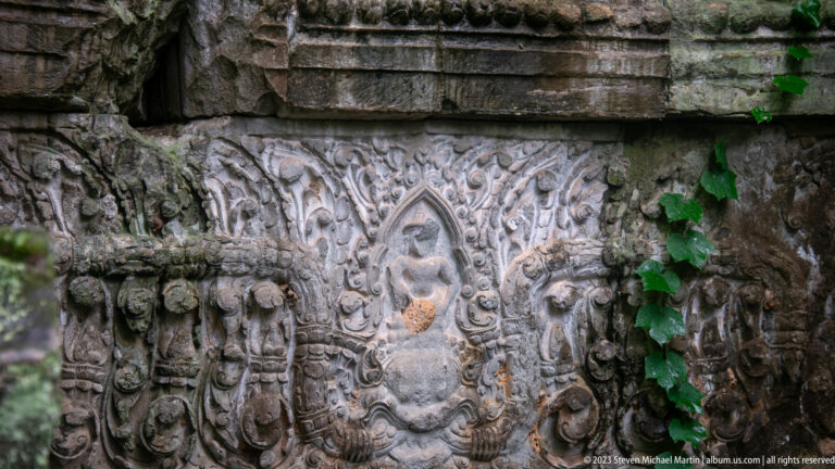 Weathered sandstone relief with a central standing figure surrounded by scrolling foliage and carved borders.