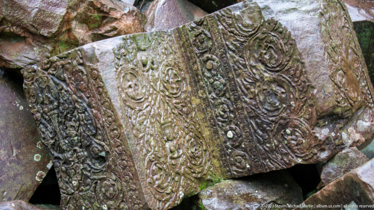 Tilted carved sandstone block with repeating foliate scrolls and beaded borders resting among temple rubble.