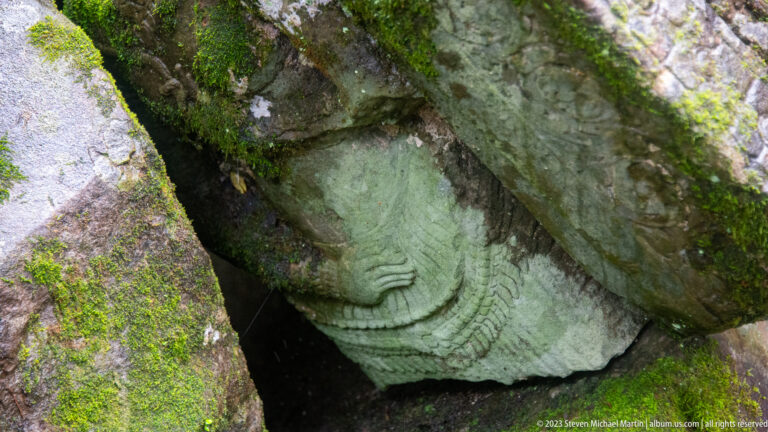 Pale carved sandstone fragment with layered curved ridges wedged beneath mossy collapsed blocks.