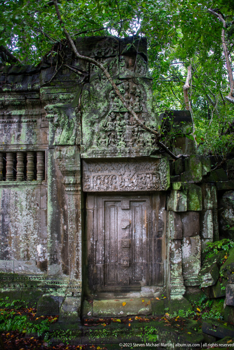Closed wooden door beneath a carved sandstone lintel, with a balustered window and rubble beside it.